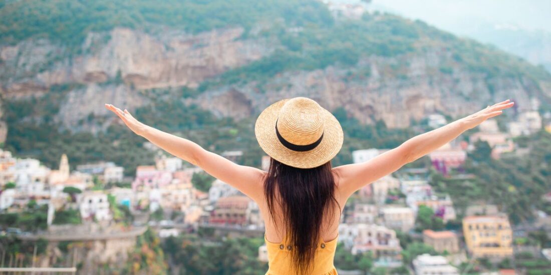 Back view of young woman in straw hat and yellow dress with Positano village on the background, Amalfi Coast, Italy