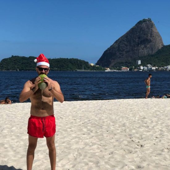 ragazzo con cappello di Natale, in costume da bagno, sulla spiaggia che beve acqua dal cocco. Mare sullo sfondo
